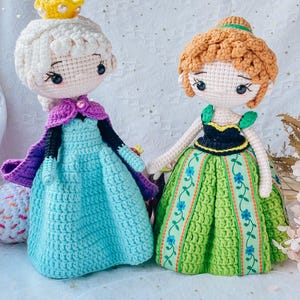 May include: Two crocheted dolls dressed as Elsa and Anna from the movie Frozen. Elsa is wearing a light blue dress with a purple cape and a gold crown. Anna is wearing a green dress with a black bodice and brown hair.