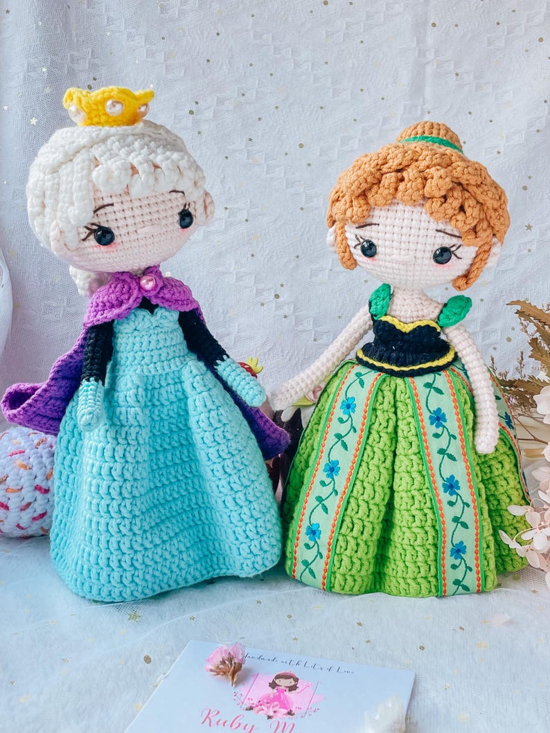 May include: Two crocheted dolls dressed as Elsa and Anna from the movie Frozen. Elsa is wearing a light blue dress with a purple cape and a gold crown. Anna is wearing a green dress with a black bodice and brown hair.