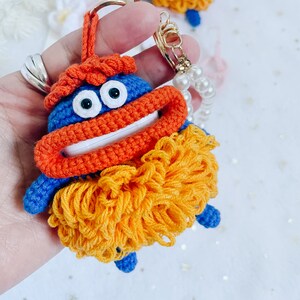 Puede incluir: Un llavero de crochet azul con hilo naranja y amarillo. El llavero tiene una cara sonriente con ojos grandes y una boca ancha. El llavero est&aacute; unido a una cadena dorada con un dije de perla.
