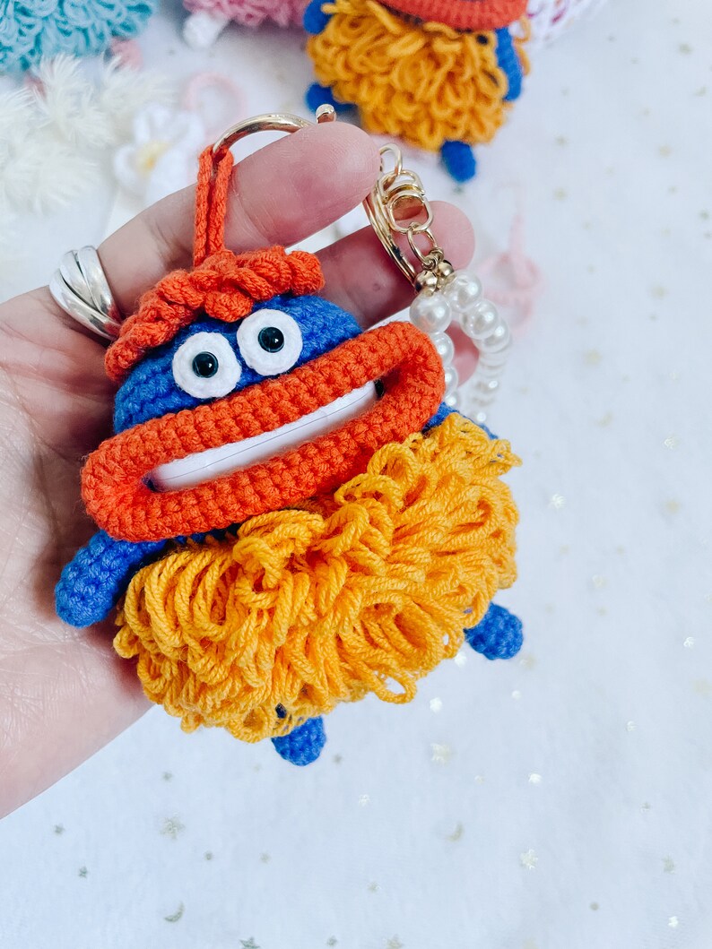 Puede incluir: Un llavero de crochet azul con hilo naranja y amarillo. El llavero tiene una cara sonriente con ojos grandes y una boca ancha. El llavero est&aacute; unido a una cadena dorada con un dije de perla.