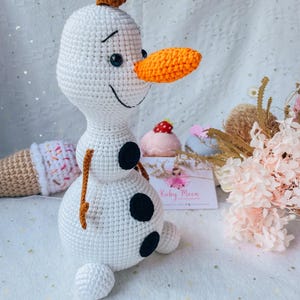 May include: A handmade crochet Olaf doll from the movie Frozen. The snowman is white with a carrot nose, black eyes, and three black buttons. It has a small brown twig for hair and arms. Other crochet items are in the background.