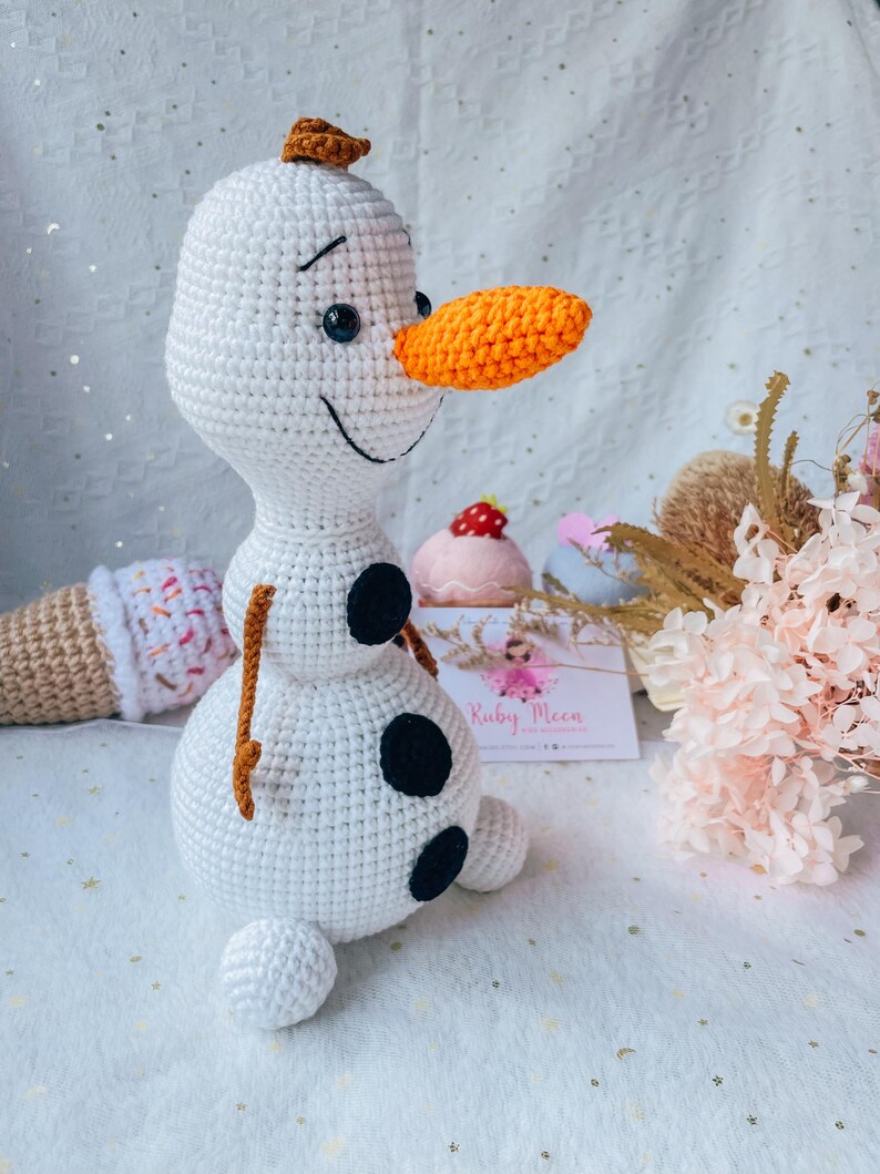 May include: A handmade crochet Olaf doll from the movie Frozen. The snowman is white with a carrot nose, black eyes, and three black buttons. It has a small brown twig for hair and arms. Other crochet items are in the background.
