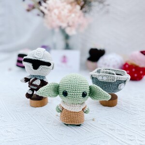 May include: Crocheted Star Wars themed figures, including a green Grogu, a Stormtrooper, and a pod. The figures are on small wooden stands. The Grogu figure has large ears and a tan outfit. The Stormtrooper has a white helmet.