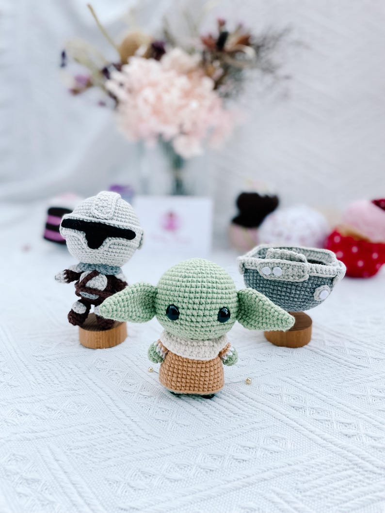 May include: Crocheted Star Wars themed figures, including a green Grogu, a Stormtrooper, and a pod. The figures are on small wooden stands. The Grogu figure has large ears and a tan outfit. The Stormtrooper has a white helmet.