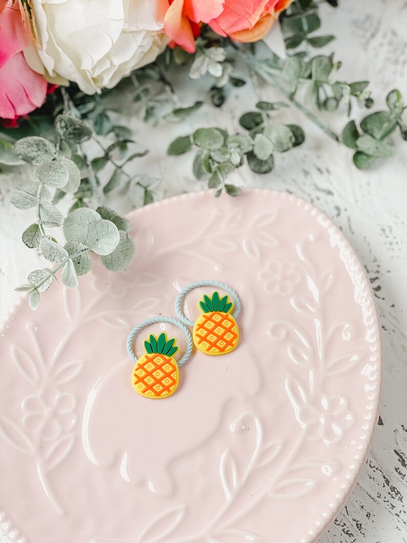 K&ouml;nnte beinhalten: Zwei blaue und gelbe Ananas-Haargummis auf einem rosa Teller mit Blumenmuster.