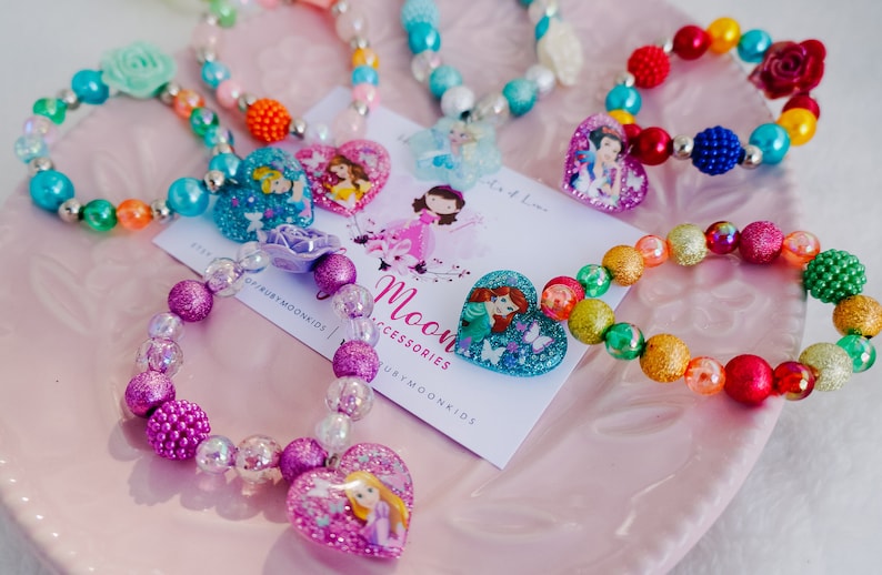 May include: A set of colorful beaded bracelets with heart-shaped charms featuring Disney princesses. The bracelets are made with various colors of beads, including pink, purple, blue, green, red, and yellow. The charms feature princesses like Ariel, Cinderella, Rapunzel, and Snow White.