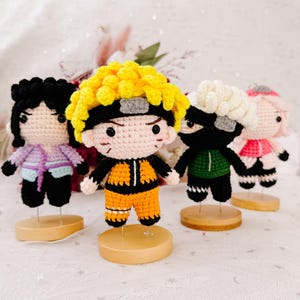 May include: Four handmade crochet dolls, each with a unique character design. The dolls feature vibrant colours, including yellow, black, orange, pink, and green. Each doll is mounted on a small wooden base, showcasing detailed craftsmanship and a playful aesthetic.