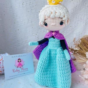 May include: Crocheted doll of a princess wearing a light blue dress, purple cape, and a yellow crown. The doll has white hair and black eyes. A small card with the text "Ruby Moon Kids Accessories" is in the background.