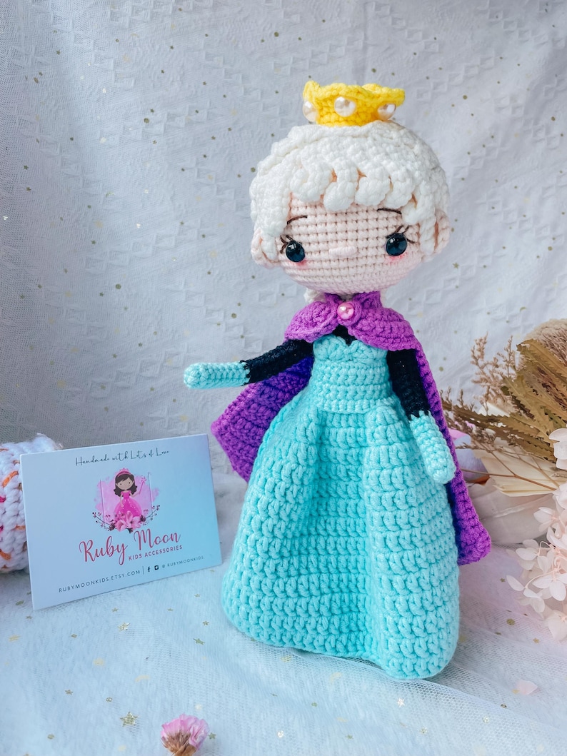 May include: Crocheted doll of a princess wearing a light blue dress, purple cape, and a yellow crown. The doll has white hair and black eyes. A small card with the text "Ruby Moon Kids Accessories" is in the background.