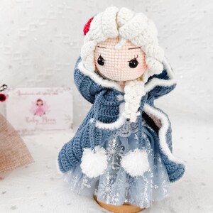 May include: A handmade crochet doll dressed in a blue and white winter outfit. The doll has a white crocheted hood with a red accent, a blue coat, and a light blue dress with snowflake details. The doll is standing on a wooden base.