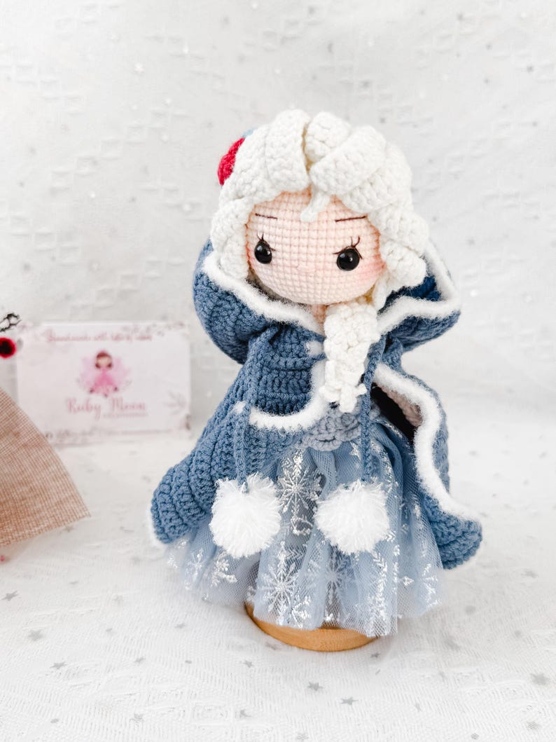 May include: A handmade crochet doll dressed in a blue and white winter outfit. The doll has a white crocheted hood with a red accent, a blue coat, and a light blue dress with snowflake details. The doll is standing on a wooden base.