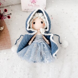 May include: A handmade crochet doll wearing a blue hooded cape with white trim and pom-poms. The doll has a light blue dress with silver snowflake accents and blonde hair. The doll is sitting on a white surface with a box in the background that says "Ruby Moon".