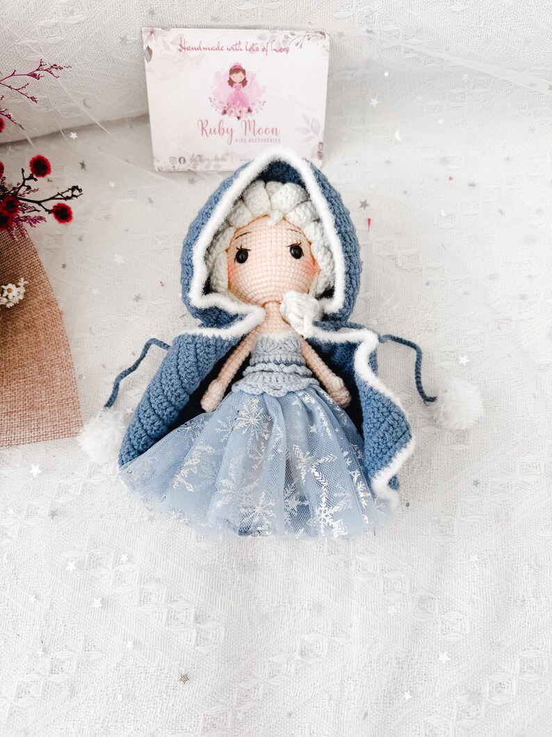 May include: A handmade crochet doll wearing a blue hooded cape with white trim and pom-poms. The doll has a light blue dress with silver snowflake accents and blonde hair. The doll is sitting on a white surface with a box in the background that says "Ruby Moon".