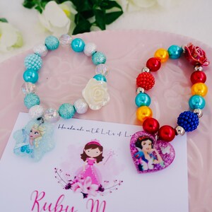 May include: Two colorful beaded bracelets with princess-themed charms. The blue bracelet has a white flower charm and a snowflake charm with a picture of Elsa from Frozen. The rainbow bracelet has a red heart charm with a picture of Snow White. The bracelets are handmade with lots of love.