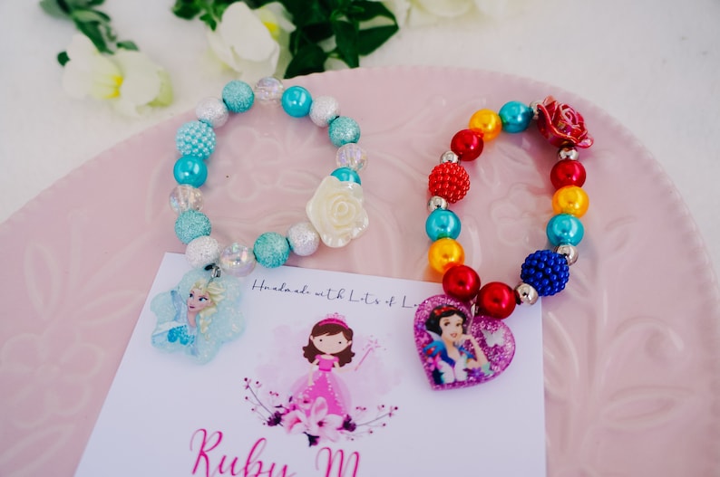 May include: Two colorful beaded bracelets with princess-themed charms. The blue bracelet has a white flower charm and a snowflake charm with a picture of Elsa from Frozen. The rainbow bracelet has a red heart charm with a picture of Snow White. The bracelets are handmade with lots of love.