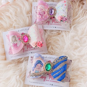 May include: Three handmade hair bows featuring Disney princesses. The bows are made with glitter, lace, and tulle. The princesses featured are Rapunzel, Cinderella, and Jasmine. The bows are on white cards with the text "Ruby Moon Kids Accessories" and their website address.