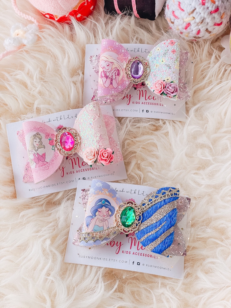 May include: Three handmade hair bows featuring Disney princesses. The bows are made with glitter, lace, and tulle. The princesses featured are Rapunzel, Cinderella, and Jasmine. The bows are on white cards with the text "Ruby Moon Kids Accessories" and their website address.