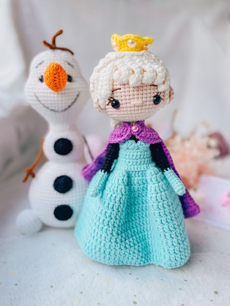 May include: Two crocheted dolls: Elsa in a blue dress with a purple cape and a yellow crown, and Olaf the snowman. The dolls are handmade with white, blue, orange, black, and brown yarn. The dolls are on a white surface.