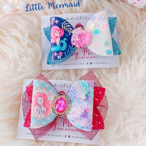 May include: Two handmade hair bows featuring a mermaid theme. The top bow is blue with white glitter and seashells, and features a mermaid with a pink tail. The bottom bow is red with a light blue and purple glitter and seashells, and features Ariel from The Little Mermaid. Both bows have the text "by Me Kids Accessories" on the card they are displayed on.