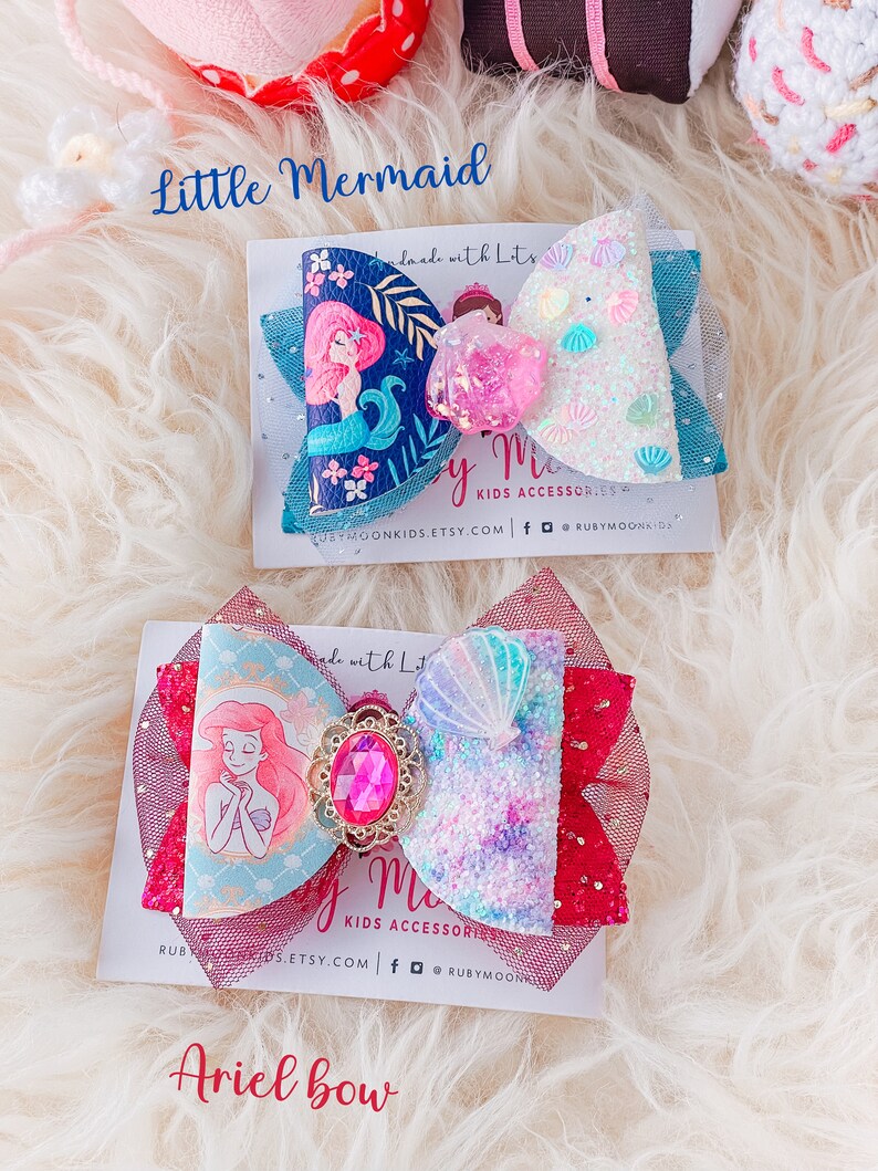 May include: Two handmade hair bows featuring a mermaid theme. The top bow is blue with white glitter and seashells, and features a mermaid with a pink tail. The bottom bow is red with a light blue and purple glitter and seashells, and features Ariel from The Little Mermaid. Both bows have the text "by Me Kids Accessories" on the card they are displayed on.