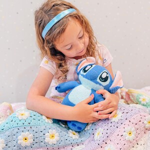 Op de afbeelding: Een jong meisje met een blauw hoofdband zit op een kleurrijk gehaakt deken met een blauw en wit pluche speelgoed. Het deken heeft een madeliefjespatroon en is gemaakt van verschillende tinten roze, paars, blauw en groen.