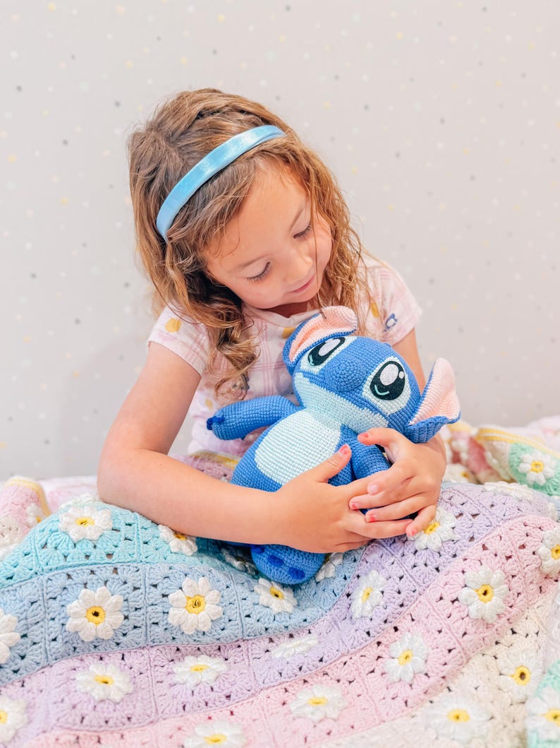 Op de afbeelding: Een jong meisje met een blauw hoofdband zit op een kleurrijk gehaakt deken met een blauw en wit pluche speelgoed. Het deken heeft een madeliefjespatroon en is gemaakt van verschillende tinten roze, paars, blauw en groen.