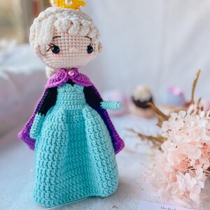 May include: A handmade crochet doll of a princess wearing a light blue dress, purple cape, and a yellow crown. The doll has white hair and is made with a tight crochet stitch. The doll is approximately 8 inches tall.