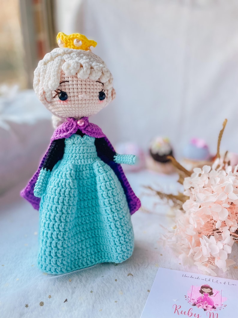 May include: A handmade crochet doll of a princess wearing a light blue dress, purple cape, and a yellow crown. The doll has white hair and is made with a tight crochet stitch. The doll is approximately 8 inches tall.