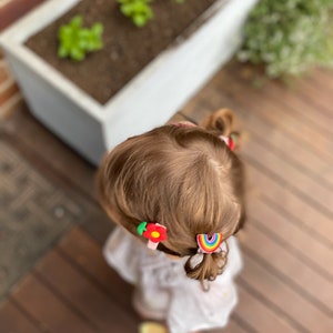 K&ouml;nnte beinhalten: Ein junges M&auml;dchen mit braunen Haaren tr&auml;gt ein wei&szlig;es Hemd und zwei bunte Haarklammern. Eine Klammer ist eine rote Blume und die andere ein Regenbogen.