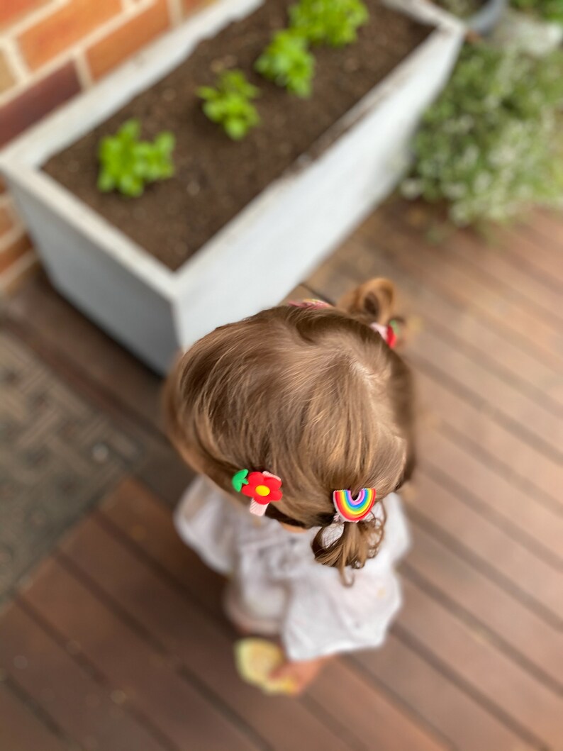 K&ouml;nnte beinhalten: Ein junges M&auml;dchen mit braunen Haaren tr&auml;gt ein wei&szlig;es Hemd und zwei bunte Haarklammern. Eine Klammer ist eine rote Blume und die andere ein Regenbogen.