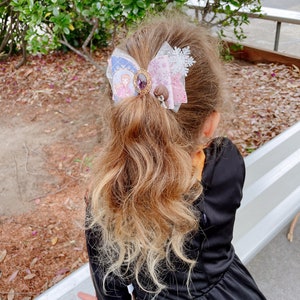May include: A young girl with long, wavy, light brown hair wearing a black dress with a large, multi-colored bow in her hair. The bow is decorated with a snowflake, a jewel, and two small figures.