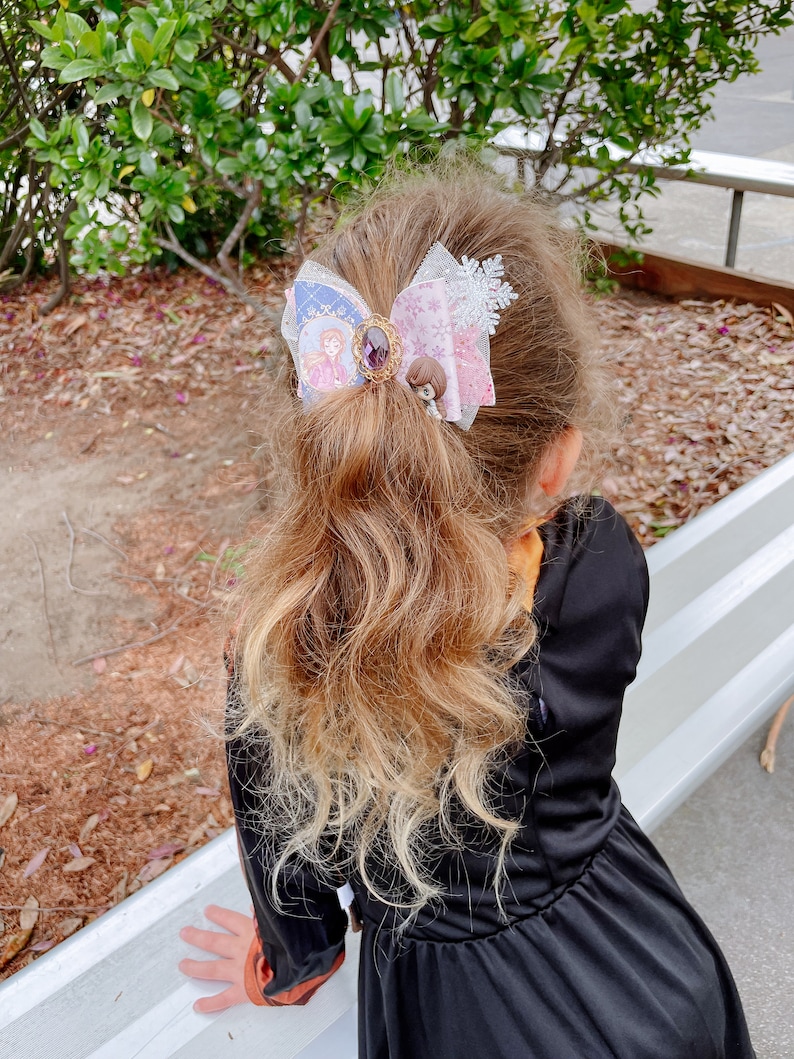 May include: A young girl with long, wavy, light brown hair wearing a black dress with a large, multi-colored bow in her hair. The bow is decorated with a snowflake, a jewel, and two small figures.