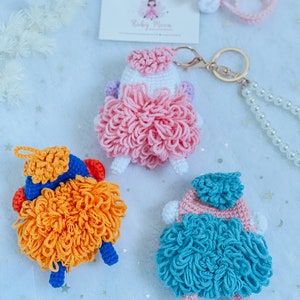 Puede incluir: Tres llaveros de oveja de crochet en rosa, naranja y azul. Cada oveja tiene un cuerpo esponjoso y una peque&ntilde;a cabeza con un gorro de crochet. Los llaveros est&aacute;n unidos a un llavero dorado.