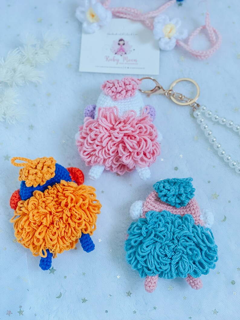 Puede incluir: Tres llaveros de oveja de crochet en rosa, naranja y azul. Cada oveja tiene un cuerpo esponjoso y una peque&ntilde;a cabeza con un gorro de crochet. Los llaveros est&aacute;n unidos a un llavero dorado.
