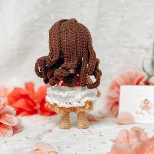 May include: A handmade crochet doll with brown hair and a white and tan skirt. The doll is wearing a red top with white polka dots. The doll is standing on a white surface with pink flowers in the background.