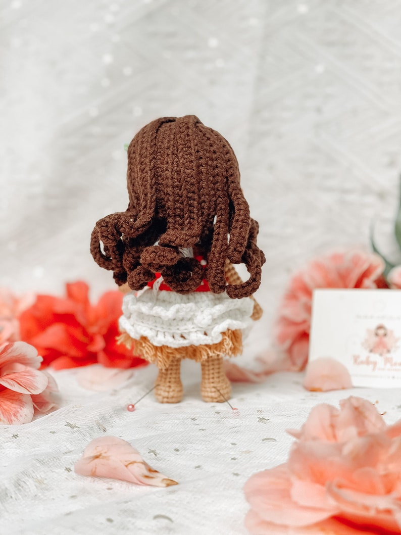May include: A handmade crochet doll with brown hair and a white and tan skirt. The doll is wearing a red top with white polka dots. The doll is standing on a white surface with pink flowers in the background.