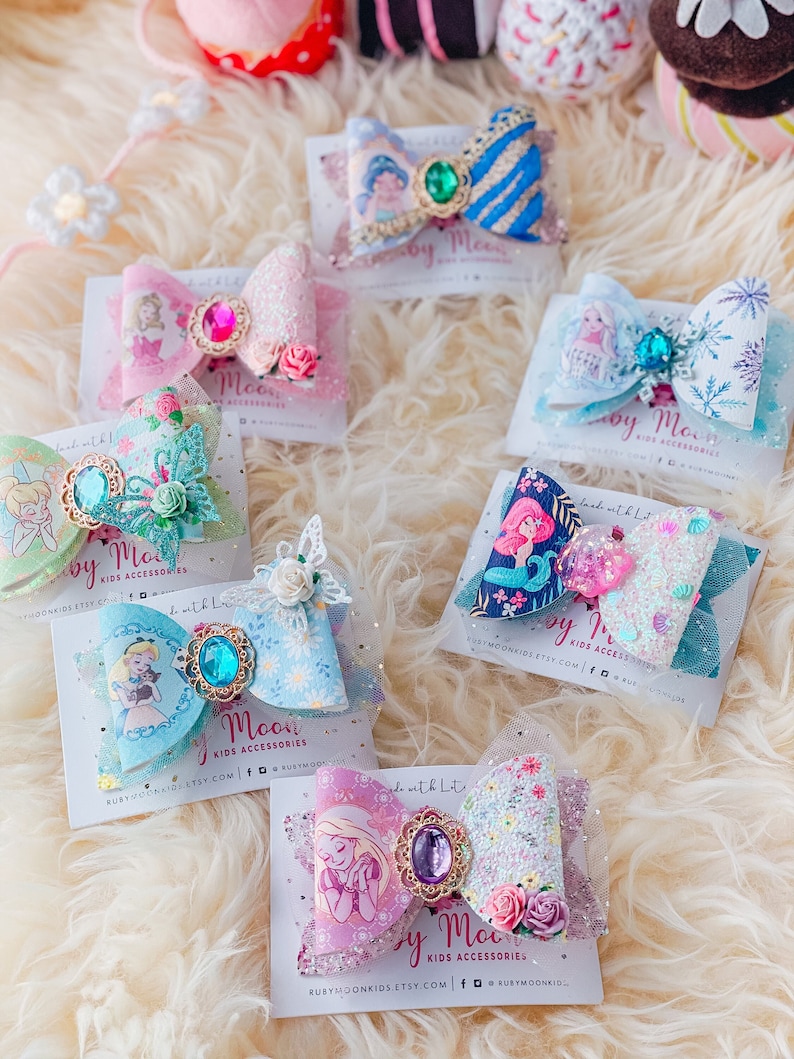 May include: A collection of handmade hair bows featuring Disney princesses. Each bow is made with glitter, tulle, and a jeweled accent. The bows are displayed on cards with the text "Ruby Moon Kids Accessories" and the website address.