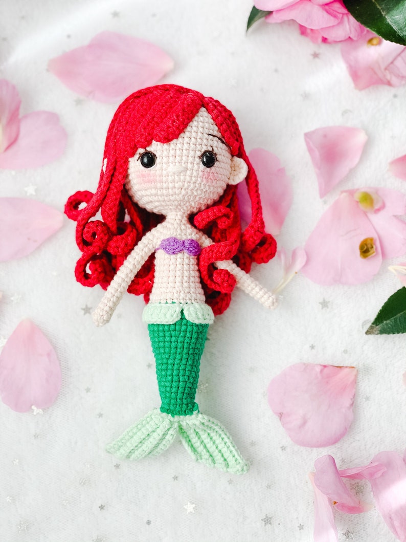 May include: A crocheted mermaid doll with red hair, a green tail, and a purple seashell top. The doll is sitting on a white background with pink flower petals.
