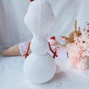 May include: A handmade crochet snowman doll with a carrot-colored hair piece and twig arms. The snowman is white with a rounded body and head. An ice cream cone and a pink dessert are in the background. The doll is a whimsical, handcrafted item.