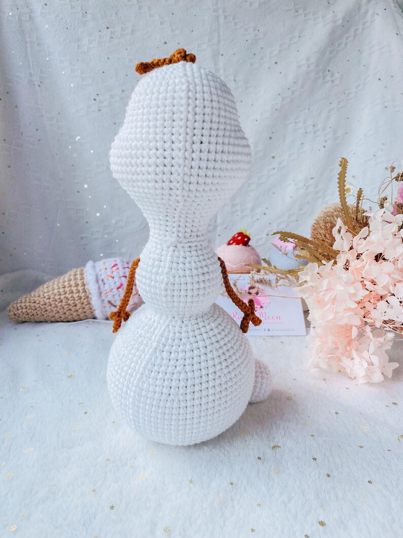May include: A handmade crochet snowman doll with a carrot-colored hair piece and twig arms. The snowman is white with a rounded body and head. An ice cream cone and a pink dessert are in the background. The doll is a whimsical, handcrafted item.
