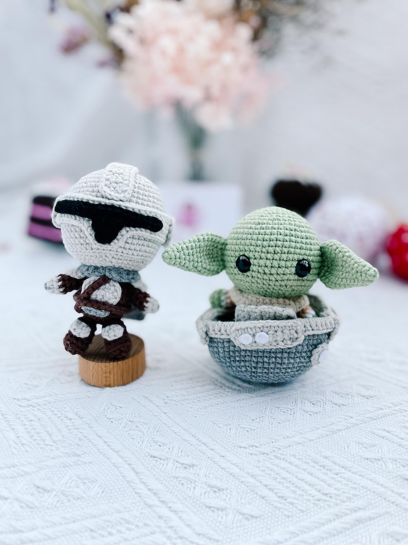 May include: Two crocheted figures: a Stormtrooper and a Grogu (Baby Yoda). The Stormtrooper is in white, gray, and brown, mounted on a wooden base. Grogu is green with large ears, seated in a gray and beige crocheted pod.