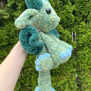 May include: A handmade, plush crochet dragon in shades of green and blue. The dragon has large, black eyes, a horn, and curled accents. The feet and accents are a lighter blue. The soft, textured yarn gives the dragon a cuddly appearance.