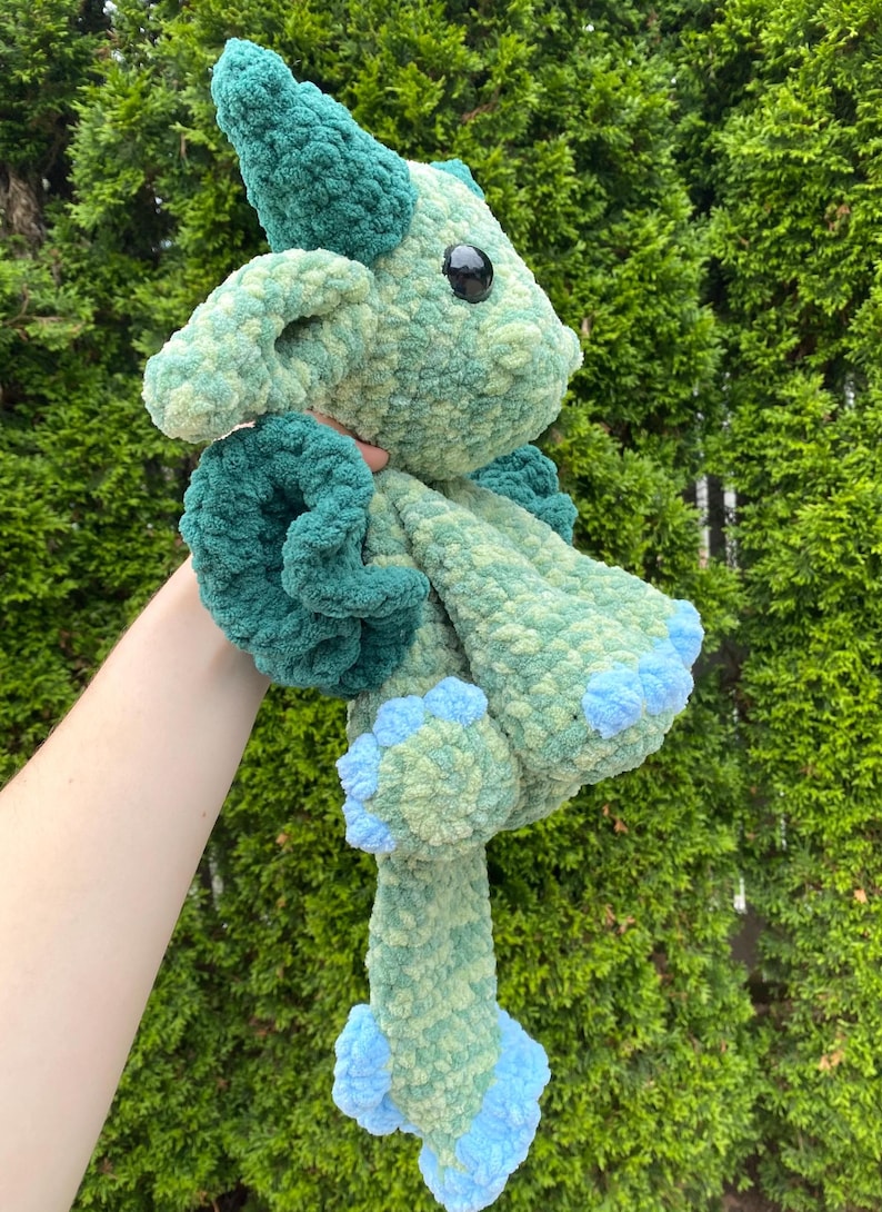 May include: A handmade, plush crochet dragon in shades of green and blue. The dragon has large, black eyes, a horn, and curled accents. The feet and accents are a lighter blue. The soft, textured yarn gives the dragon a cuddly appearance.