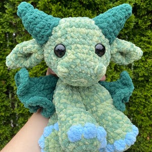 May include: A handmade, plush dragon toy in shades of green and blue. The dragon has large, black eyes, horns, wings, and blue accents on its feet. The soft, textured material gives the dragon a cuddly appearance.