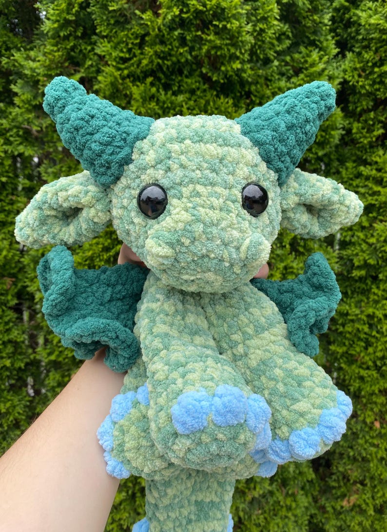 May include: A handmade, plush dragon toy in shades of green and blue. The dragon has large, black eyes, horns, wings, and blue accents on its feet. The soft, textured material gives the dragon a cuddly appearance.
