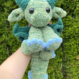 May include: A handmade, plush dragon toy in shades of green and blue. The dragon has large black eyes, horns, wings, and blue accents on its feet and tail. The soft, textured material gives it a cuddly appearance.