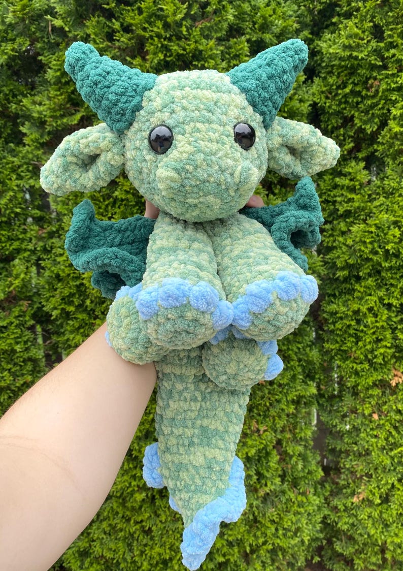 May include: A handmade, plush dragon toy in shades of green and blue. The dragon has large black eyes, horns, wings, and blue accents on its feet and tail. The soft, textured material gives it a cuddly appearance.