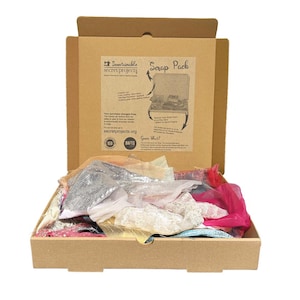 May include: A brown cardboard box filled with assorted fabric scraps in various colours and textures. The inside lid features a printed label with the text 'Sustainable Secret Projects' and 'Scrap Pack' along with descriptive information and logos.