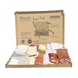 May include: A brown cardboard box labelled Sewtainable Secret Projects Scrap Pack Bridal containing an assortment of folded fabric scraps in various textures and colours including white lace, pink satin, and a floral patterned piece of fabric.
