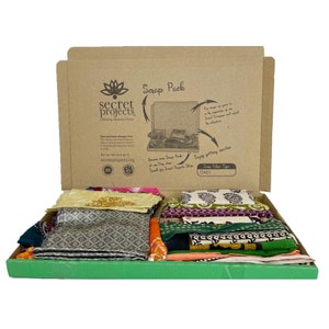 May include: An open cardboard box with a green lid, filled with colourful fabric scraps. The box is labelled "Scrap Pack" and features the logo for "Secret Projects: Unlocking Women's Power".